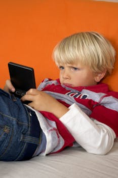 Blond boy playing on handheld console while lounging on a sofa indoors.