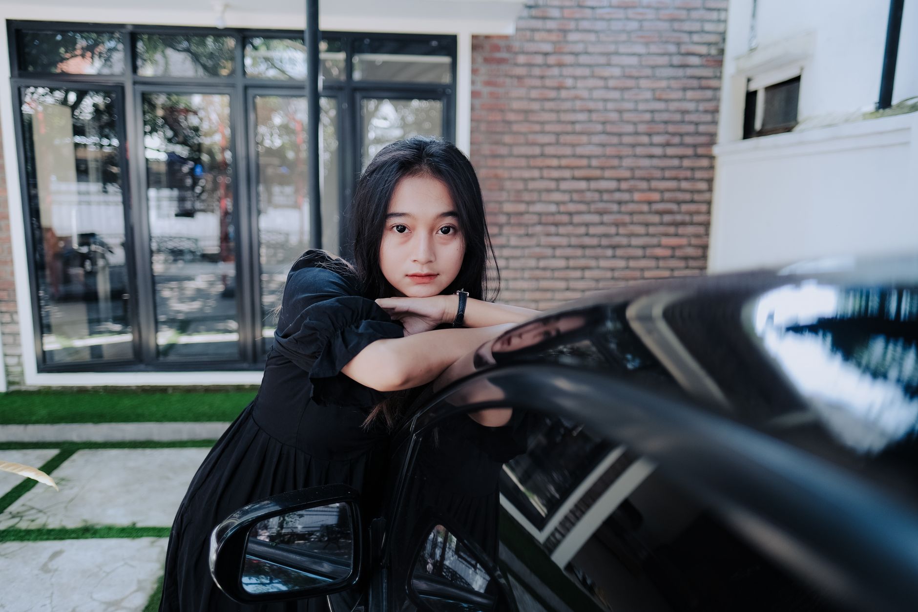 A young woman in a black dress leans against a car in an outdoor setting.