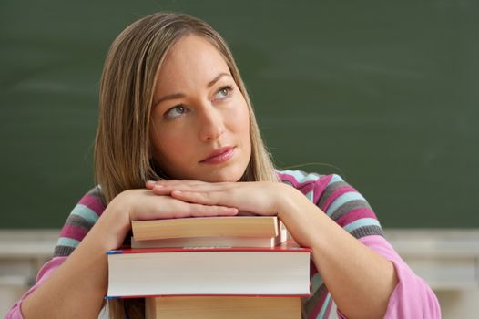 Young woman in a classroom, leaning on a stack of books, deep in thought.