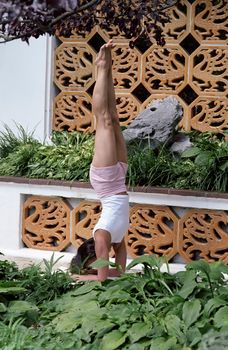 Woman practicing a yoga headstand in a lush garden with a decorative wall.