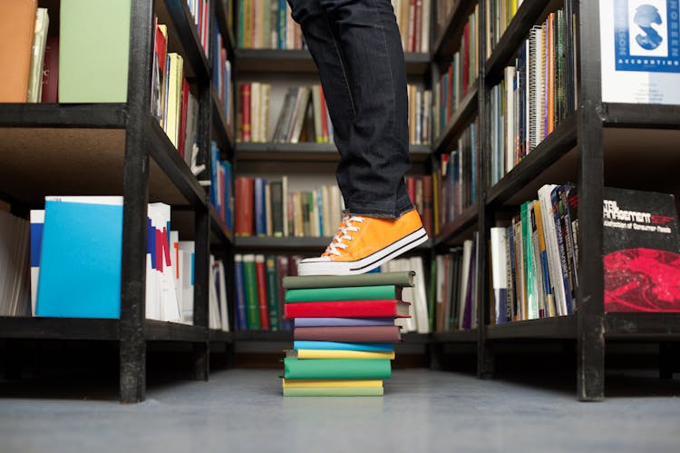 Leg Of A Person Standing On Top Of A Stack Of Books In A Library