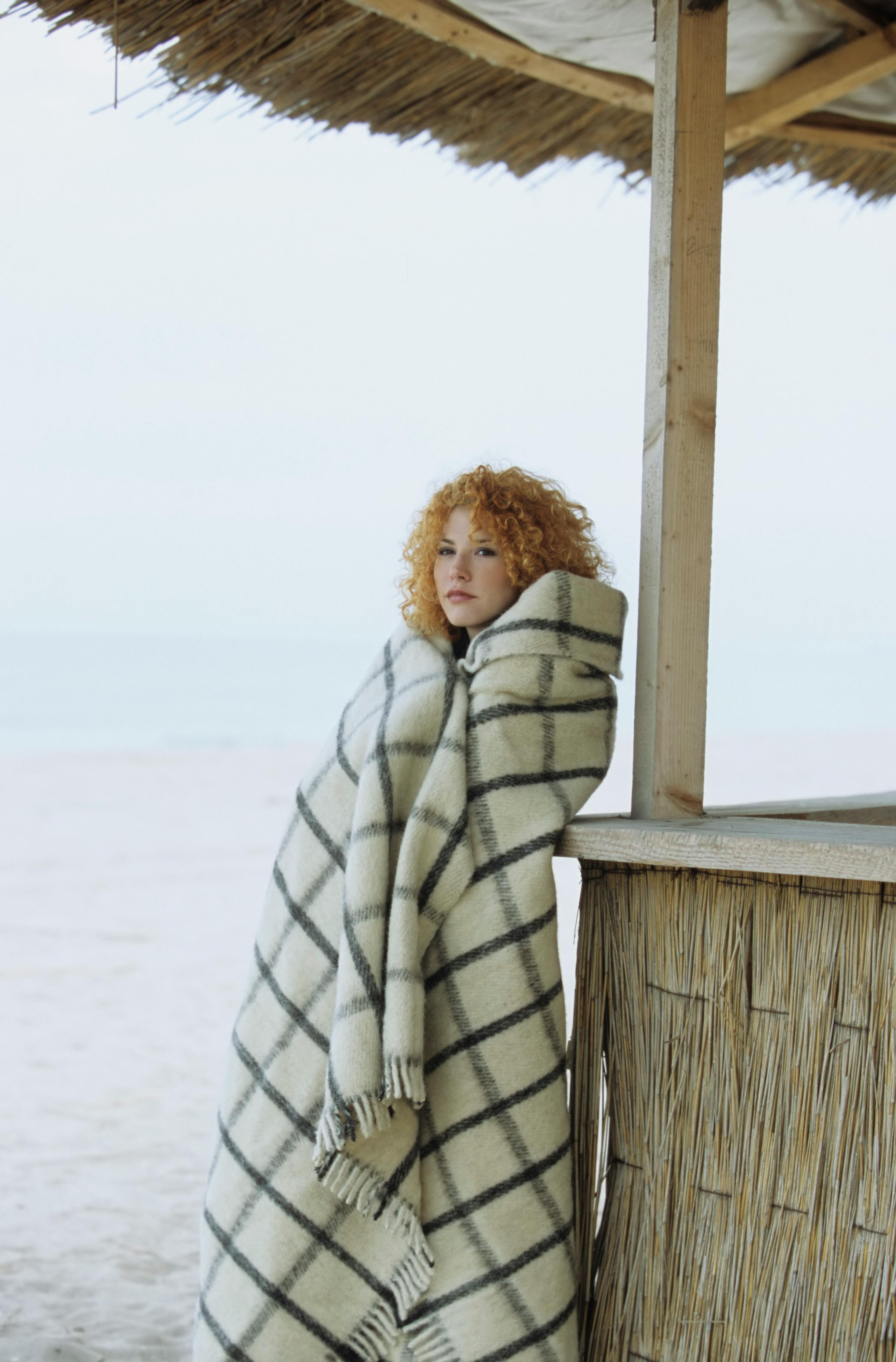 Woman wrapped in a checkered blanket standing by a beach hut on a cool day.