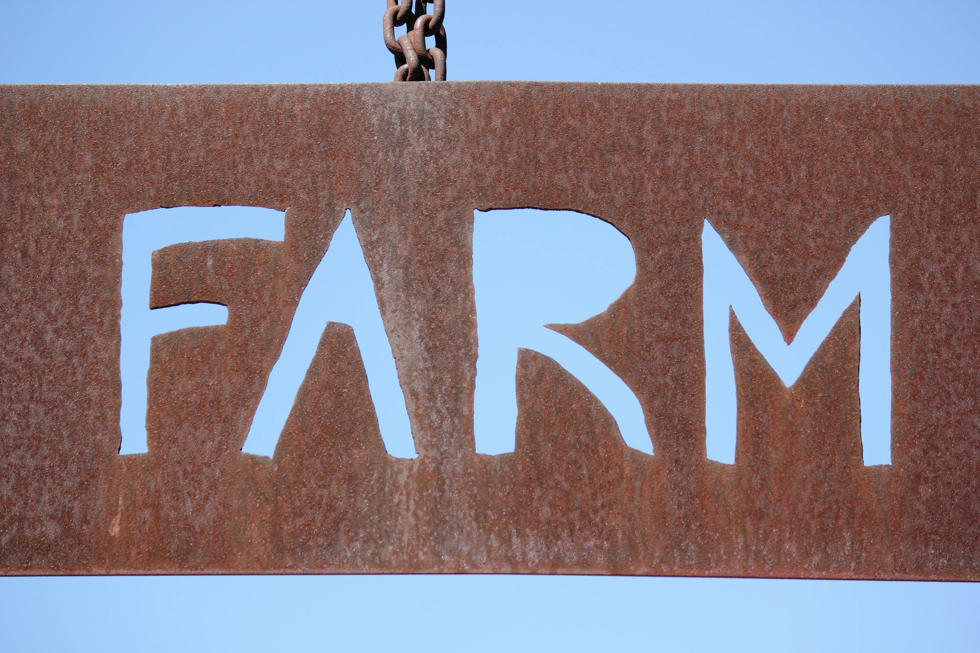 Close-up of a rusty metal farm sign with cut-out letters against a blue sky