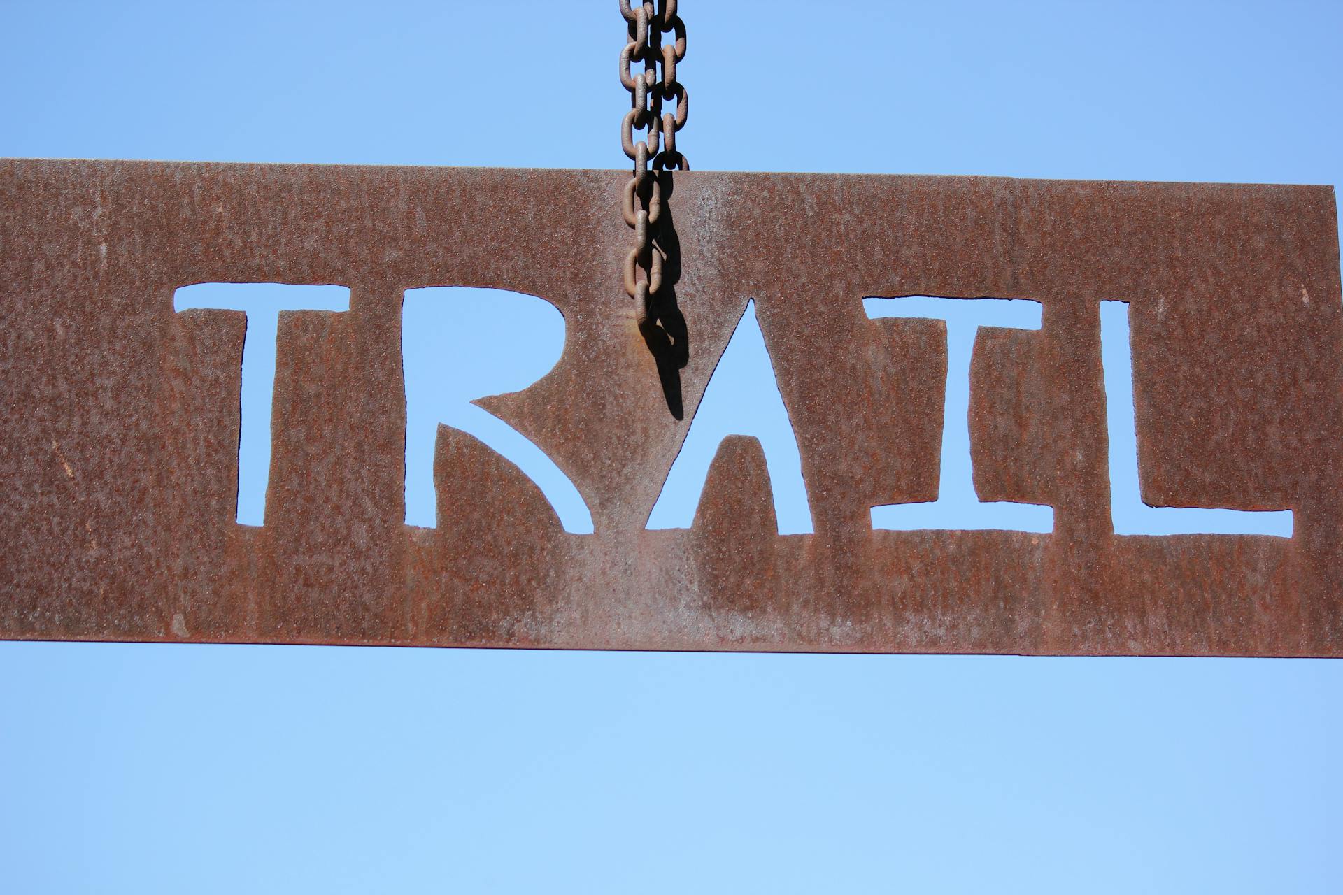 Close-up of a rusty trail sign hanging by a chain against a clear blue sky.