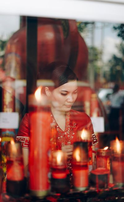An adult Asian woman in traditional attire reflects behind a glass panel with candles in a ritual setting.