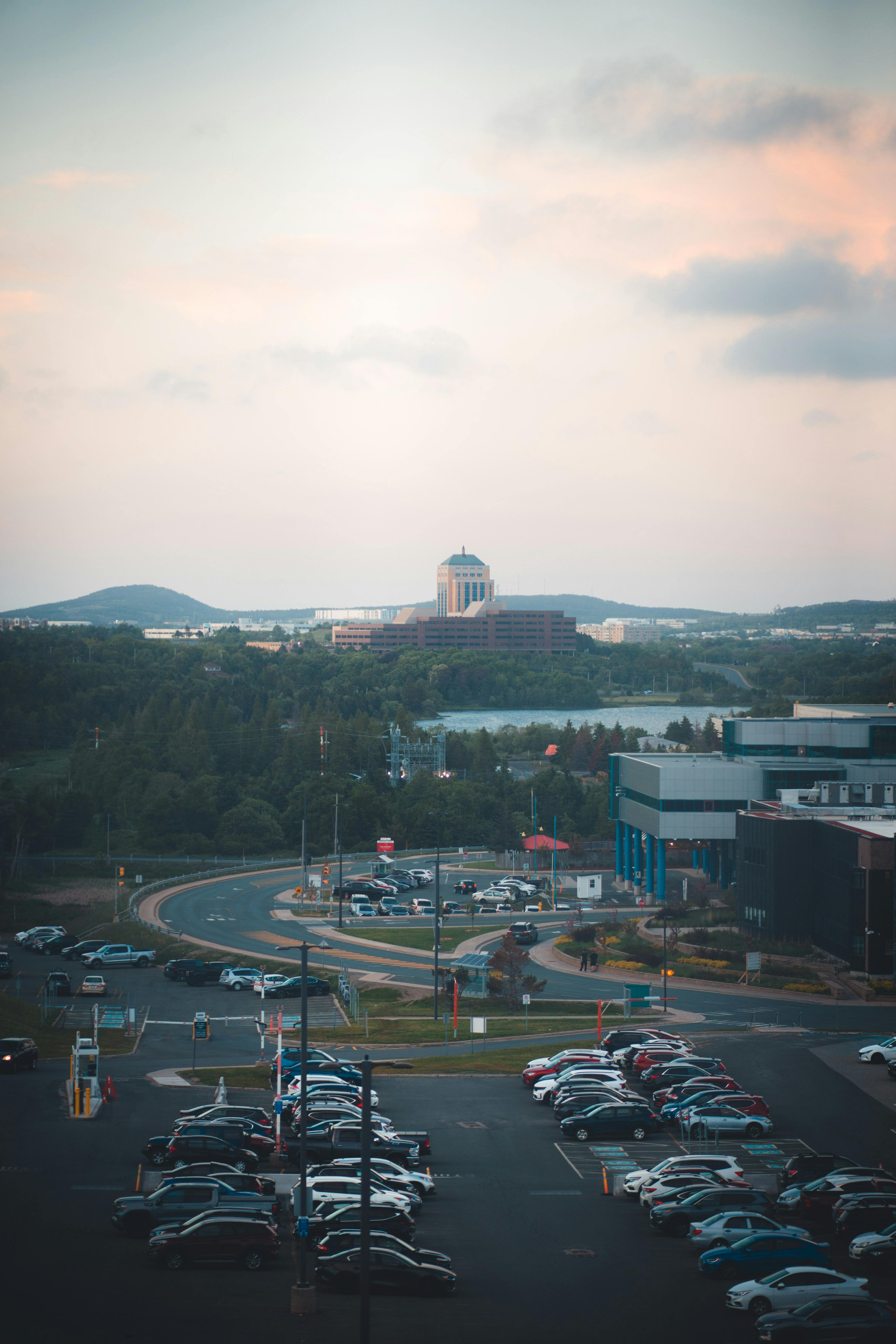 Aerial urban view featuring a car park, buildings, and surrounding natural scenery under a soft sky.