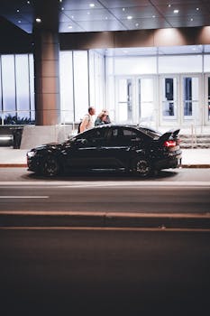 Side view of a black car parked on a city street at night with people walking nearby.