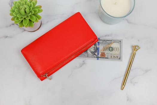 A chic red wallet with a candle, dollar bill, golden pen, and succulent on a marble surface.