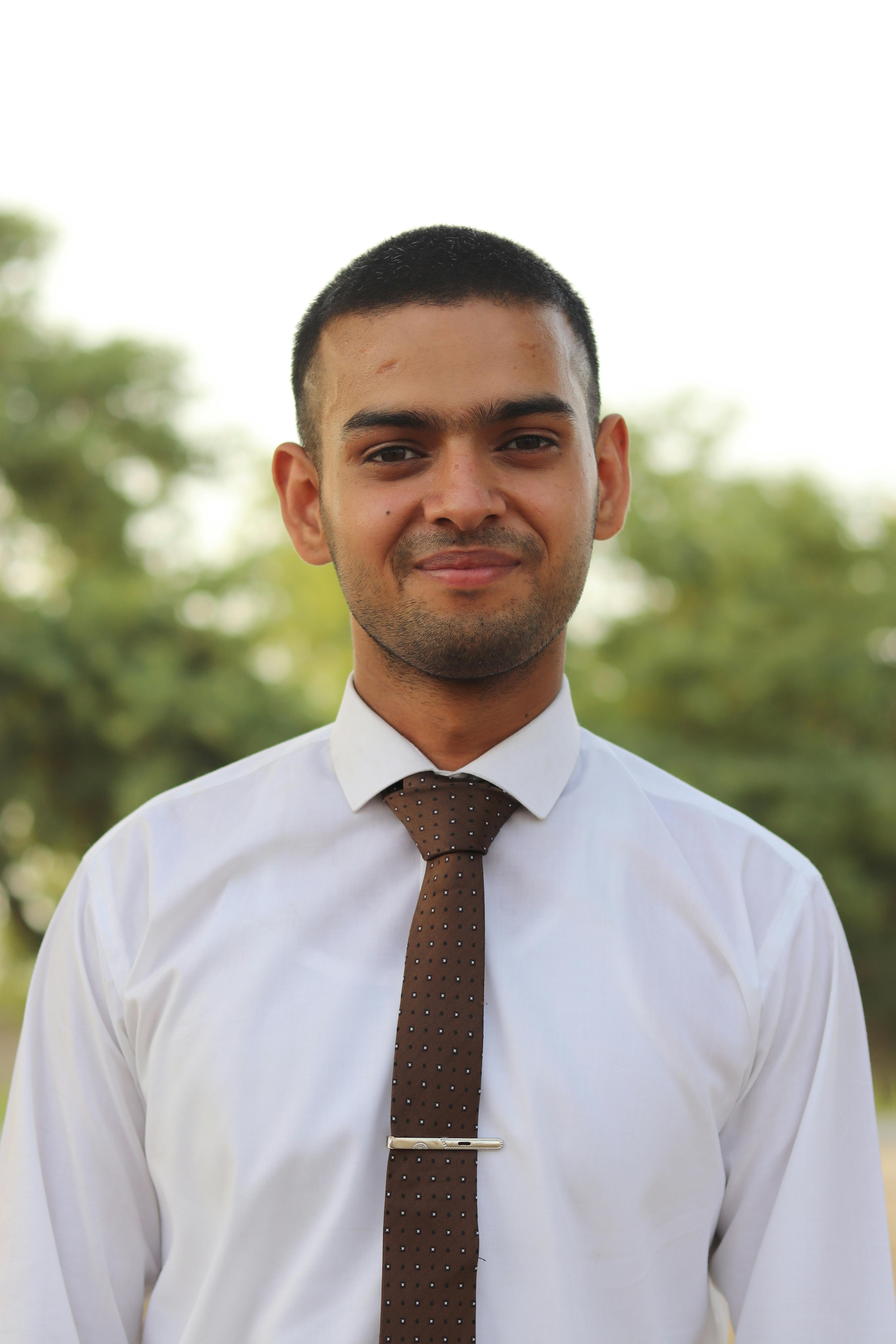 A confident young man in a white shirt and necktie poses outdoors with a warm smile.