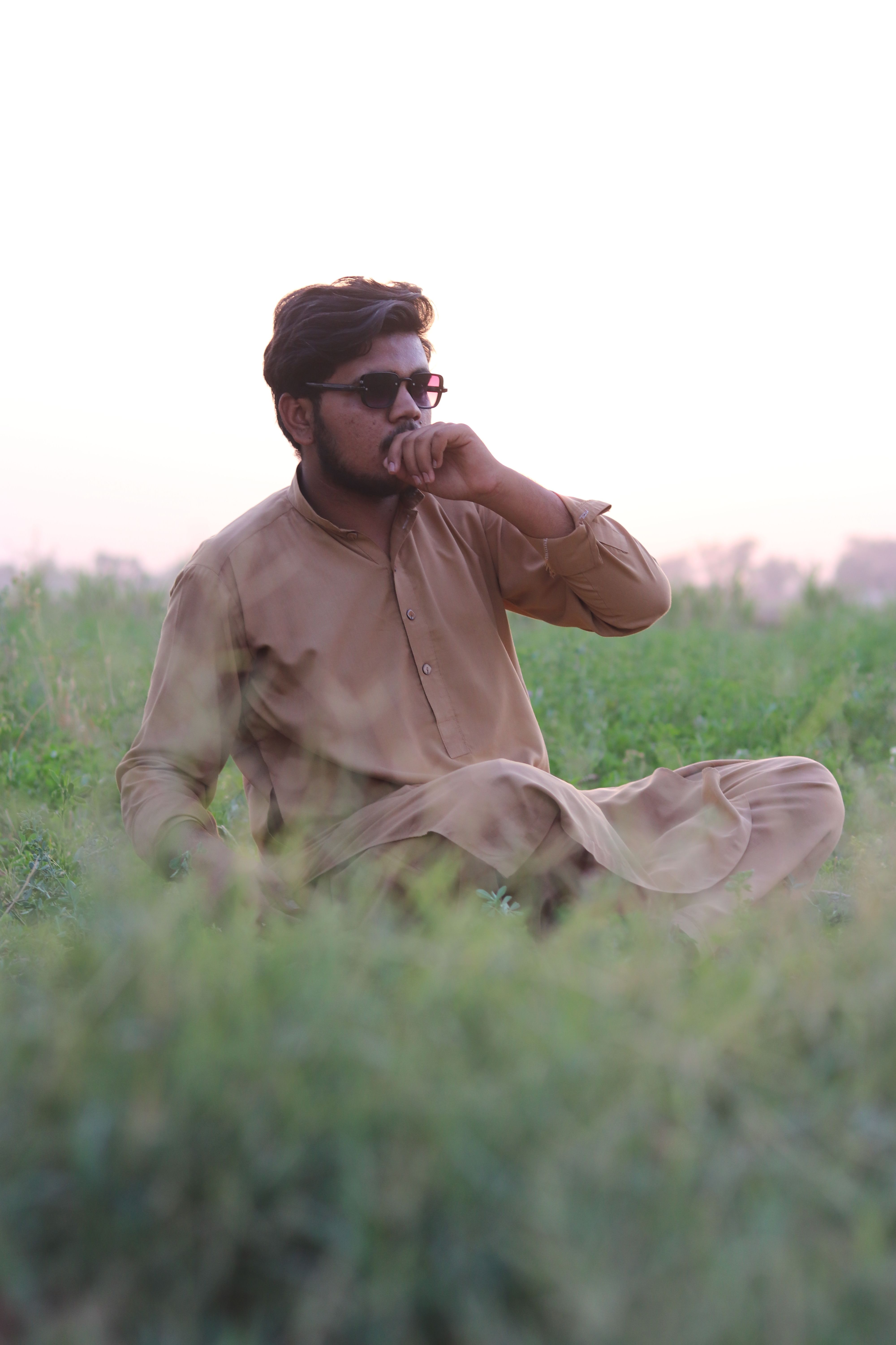 A man wearing sunglasses and traditional attire sits in a lush green field during sunset.