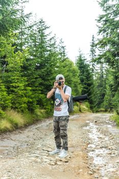 A young man with a camera takes photos on a forest trail surrounded by lush trees.