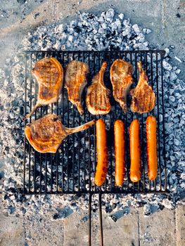 Top view of grilled pork chops and sausages over charcoal ashes.