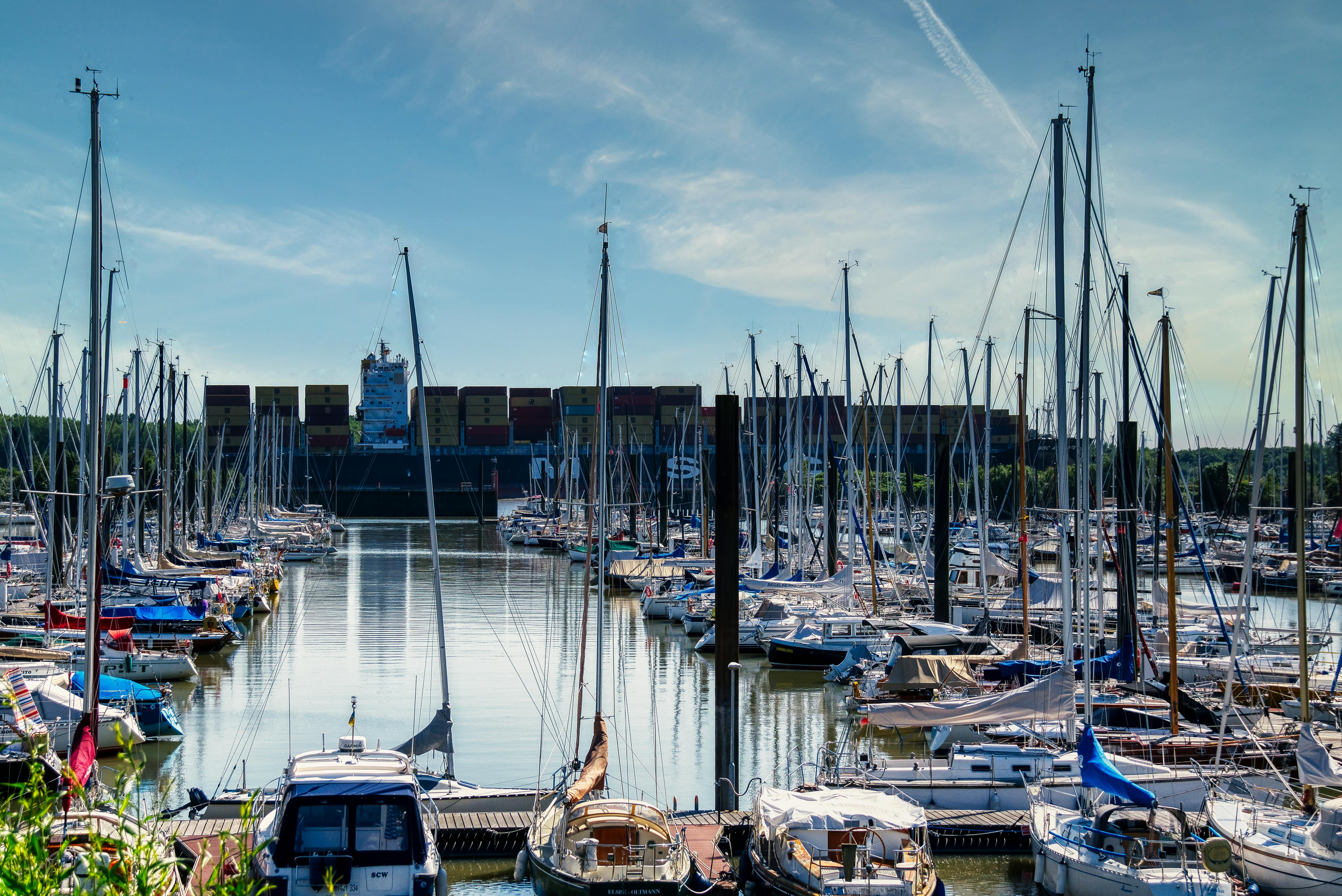 A vibrant marina with moored yachts and a distant view of a container ship under a bright sky.