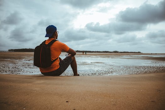 Man wearing a backpack sitting on a quiet beach, contemplating the serene seascape under a cloudy sky.