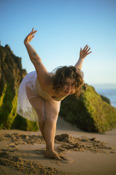 Woman practicing yoga on sandy beach with rock formations at sunset.