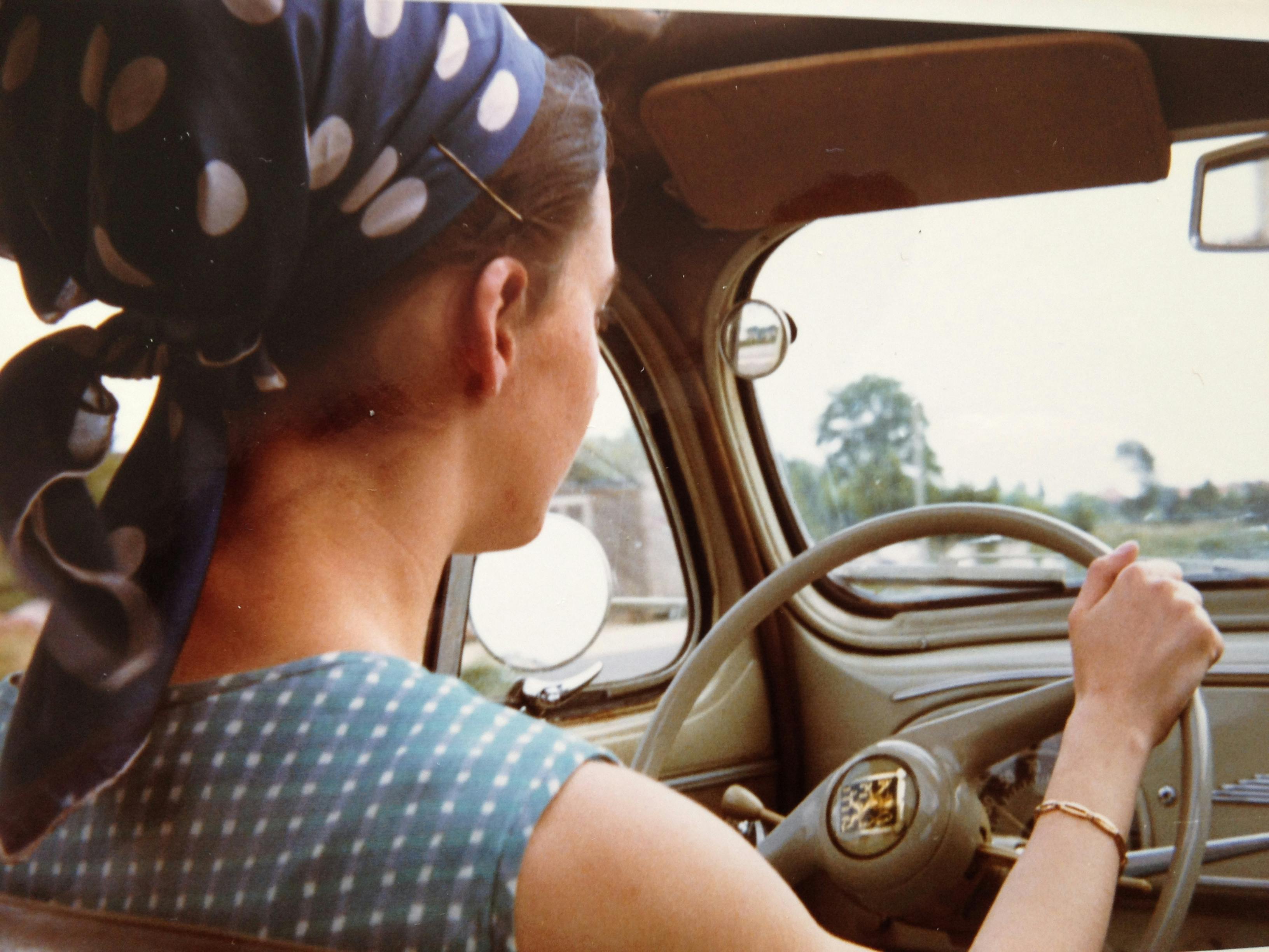 A woman wearing a headscarf drives a classic vintage car, evoking a retro vibe.