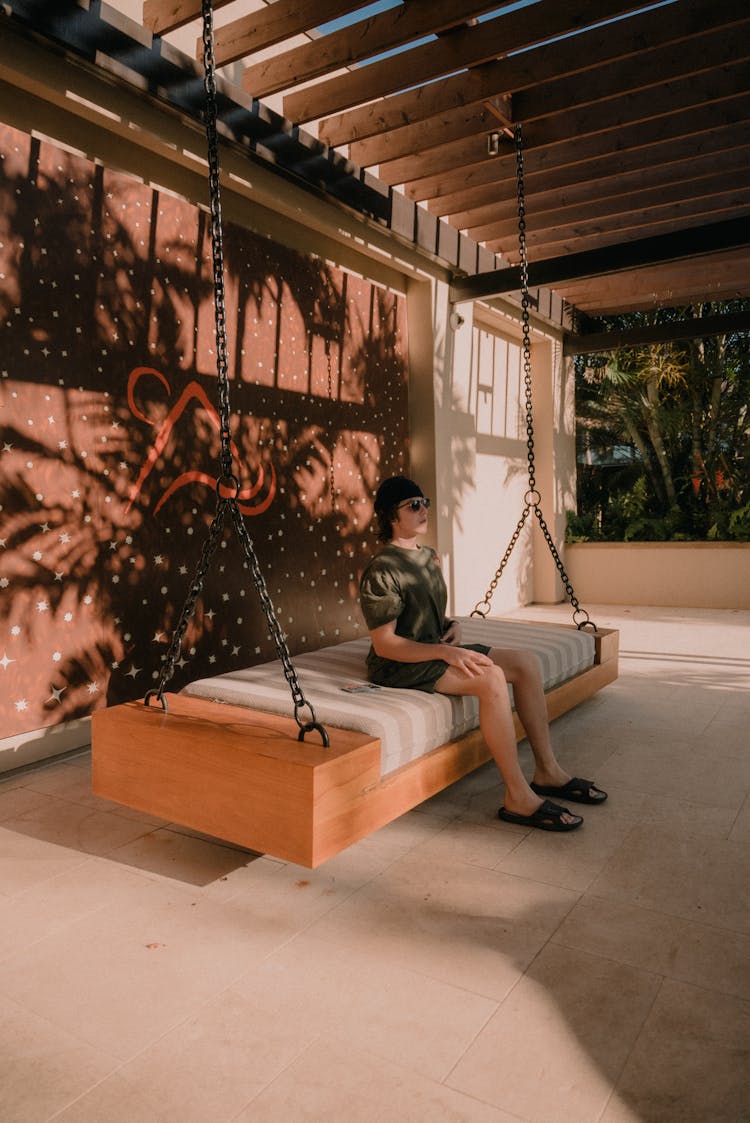 Young Man Sitting On A Hanging Bed On A Patio In A Resort 