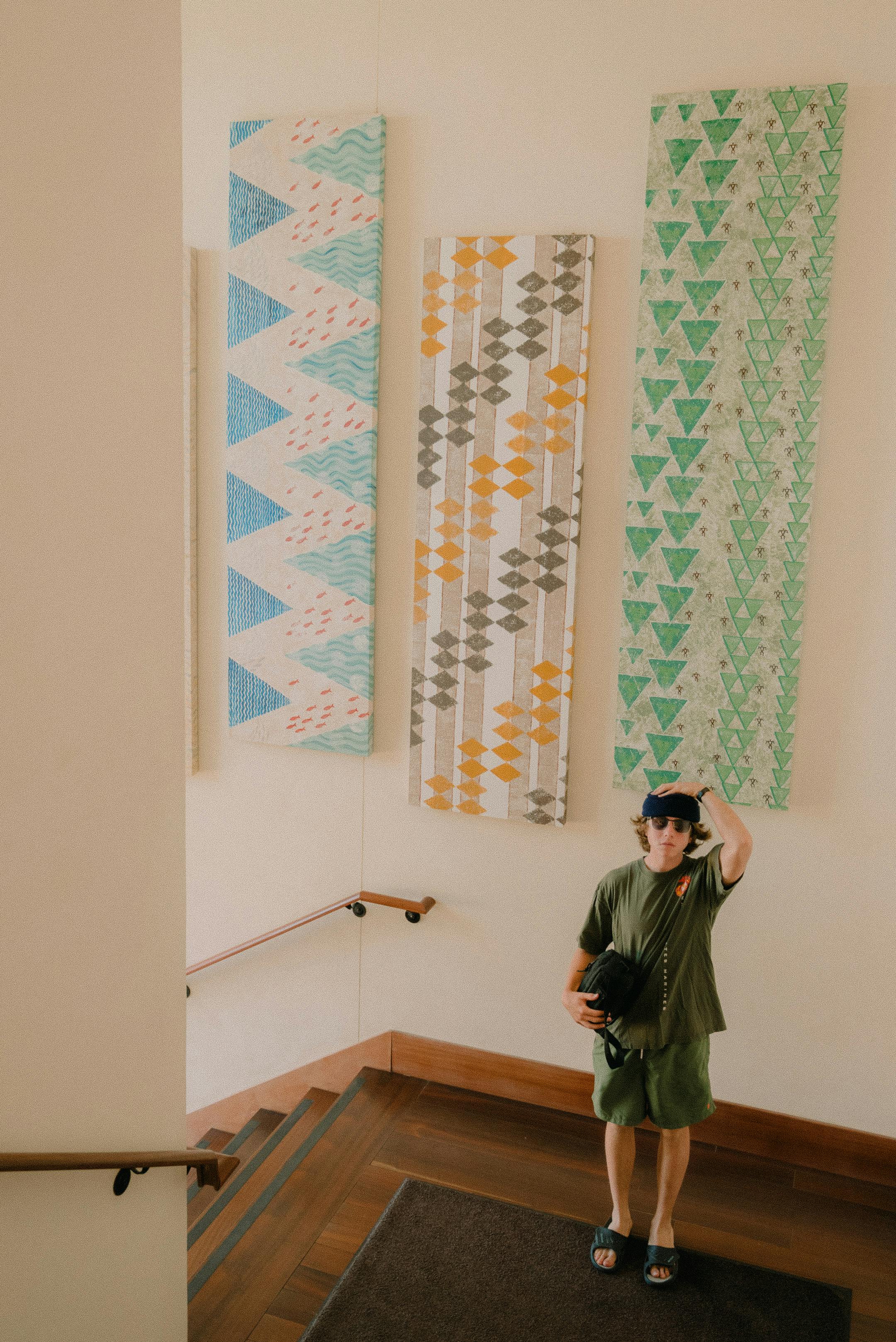 Adult man in a green outfit wearing sunglasses, standing near a colorful wall art in a stairwell.