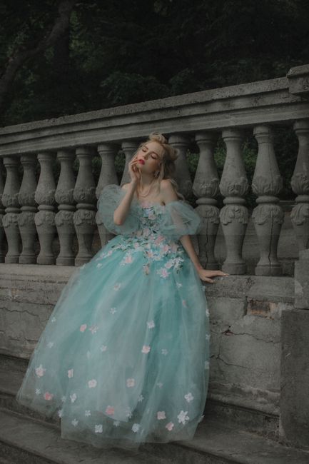 A woman in a floral gown poses elegantly by a stone railing outdoors.