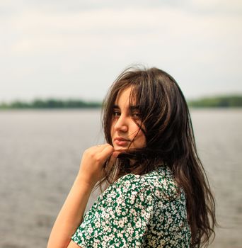 A young woman gazes thoughtfully at the camera beside a serene lake on a calm day.