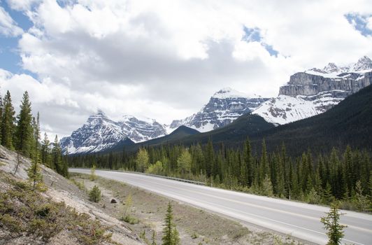 Stunning view of a mountain road surrounded by pine trees and snow-covered peaks under a cloudy sky.