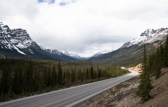 Explore a winding mountain road in a forested valley with snow-capped peaks under a cloudy sky.