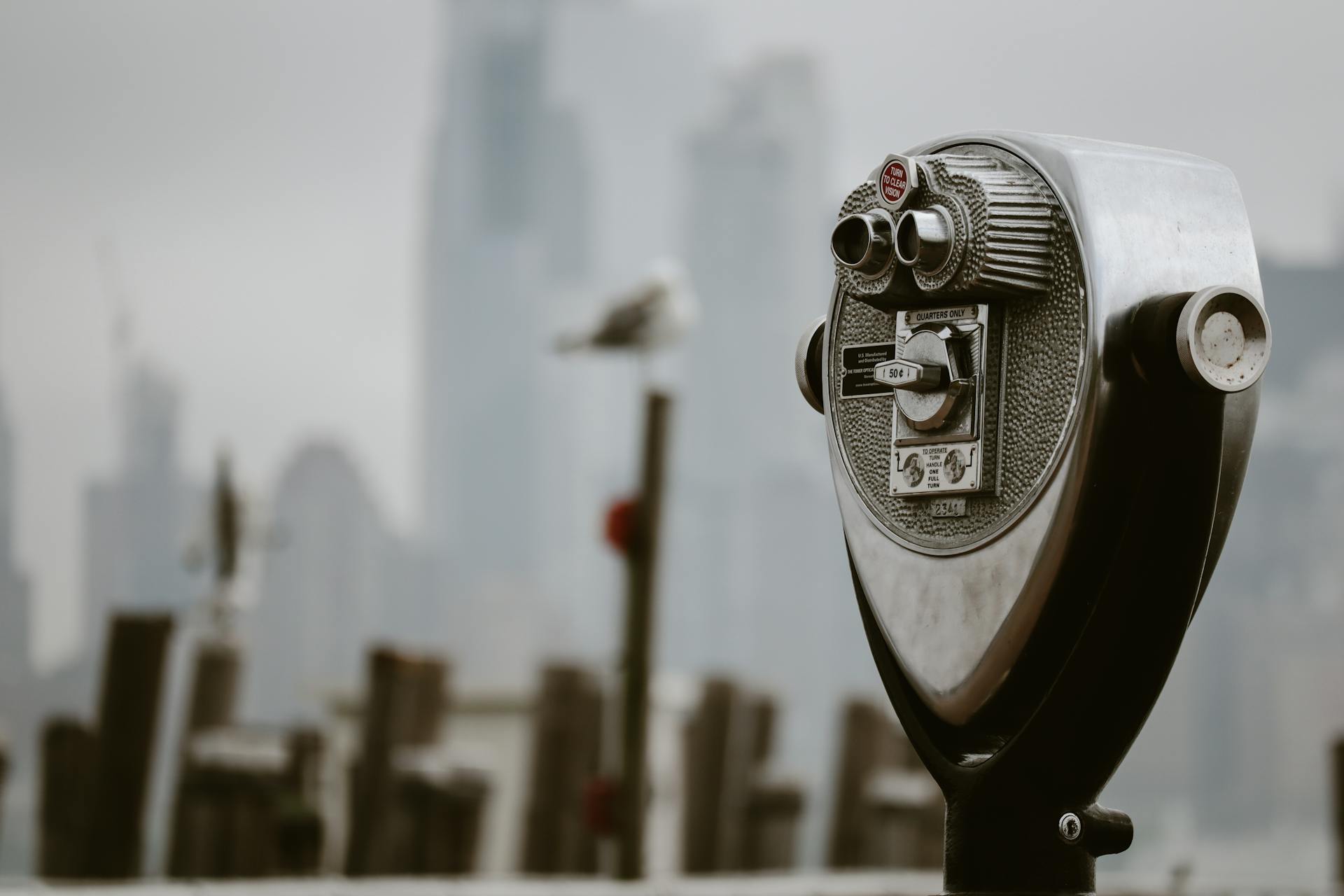 A coin-operated binocular viewer overlooks a blurred New York City skyline on a cloudy day.