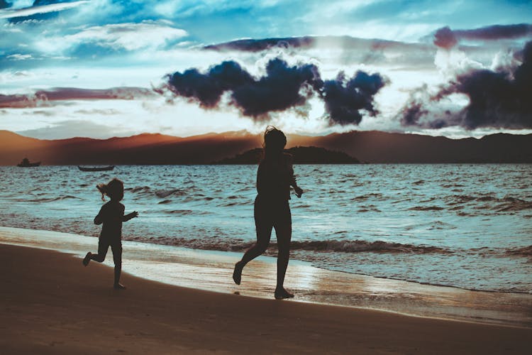 A Silhouette Of A Mother And Daughter Running On A Beach