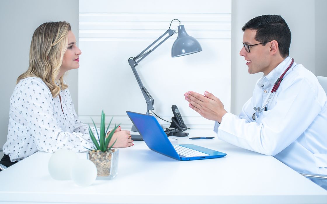A woman talks with a doctor across a desk during a consultation in a bright medical office.
