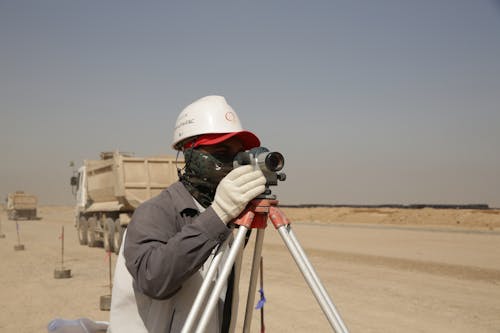 Anonymous male geodesist in protective helmet and gloves using tripod with tacheometer while determining territory frontiers at work standing on sandy road near trucks