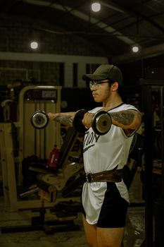 Muscular tattooed man with glasses lifting dumbbells indoors in a gym.