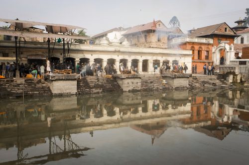 Free A serene reflection of Pashupatinath Temple's cremation ghats on the Bagmati River with people gathered. Stock Photo