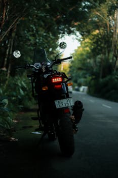 A motorcycle parked on a quiet, scenic road in KL, India. Lush greenery surrounds the area, providing a tranquil setting.