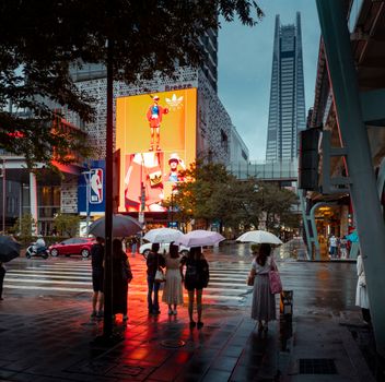 People with umbrellas crossing a wet street in downtown Taipei, with illuminated billboards and skyscrapers in the background.
