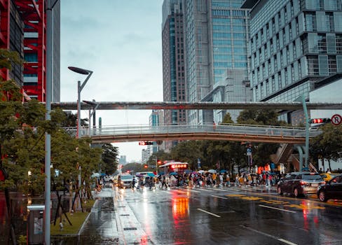 A vibrant city scene during a rainy evening showcasing a pedestrian bridge and busy street.