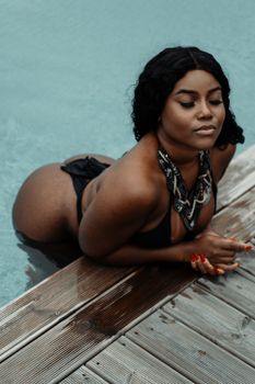 A woman in a black bikini enjoys a calm moment on a wooden poolside deck.