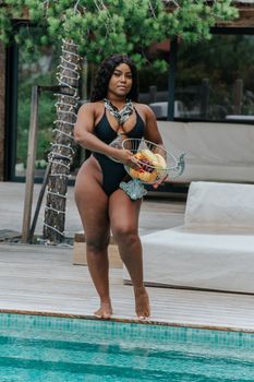 Fashionable woman in swimwear holds a fruit basket by the poolside on a warm summer day.