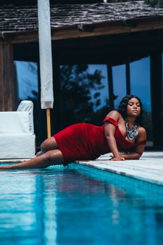 A stylish woman in a red dress poses gracefully by the swimming pool, exuding elegance.