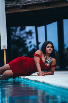 Stylish woman in red dress posing elegantly by a poolside, showcasing fashion and beauty.
