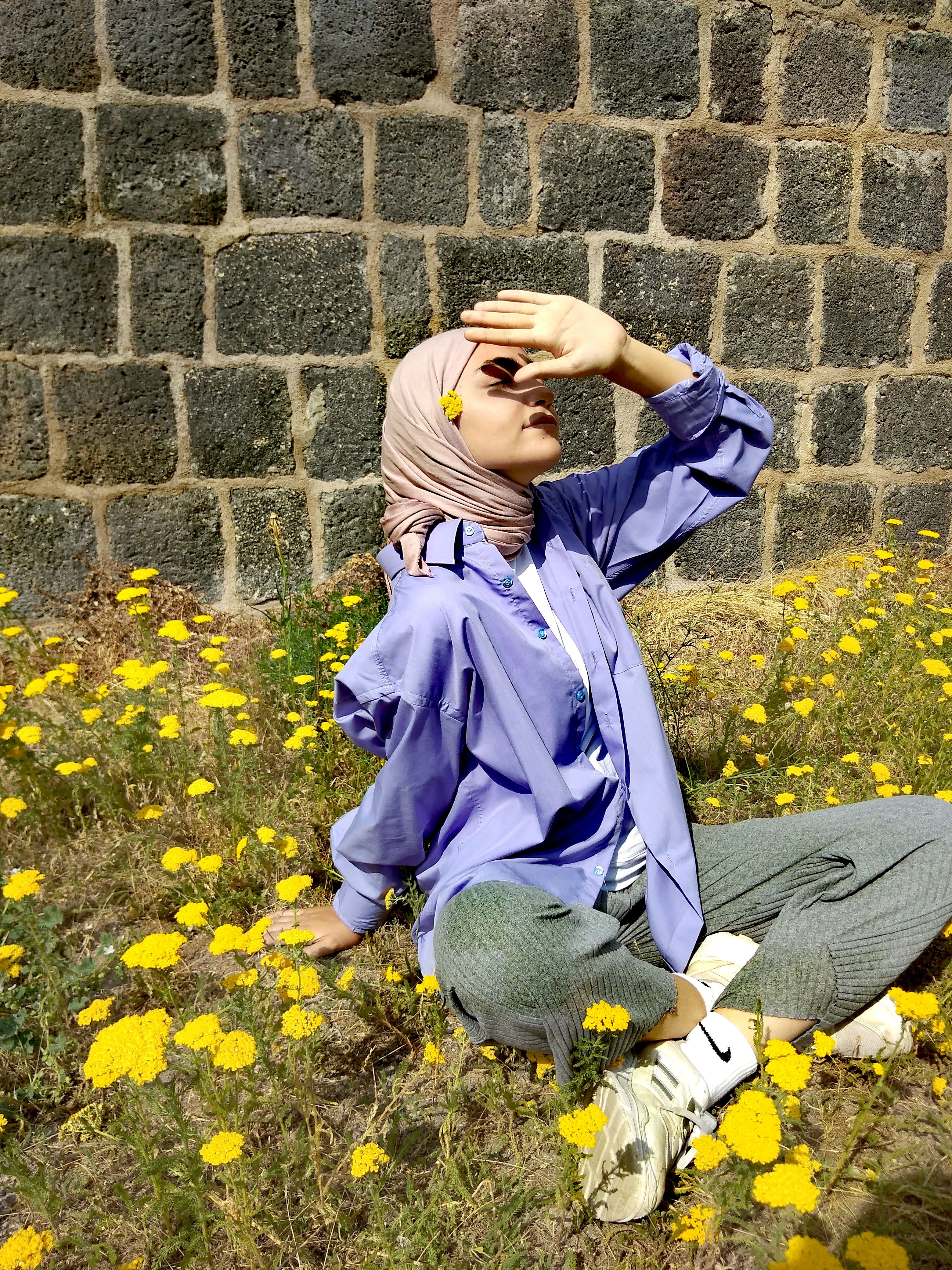 A woman in a hijab sits among vibrant yellow flowers under sunlight, smiling in a serene outdoor setting.