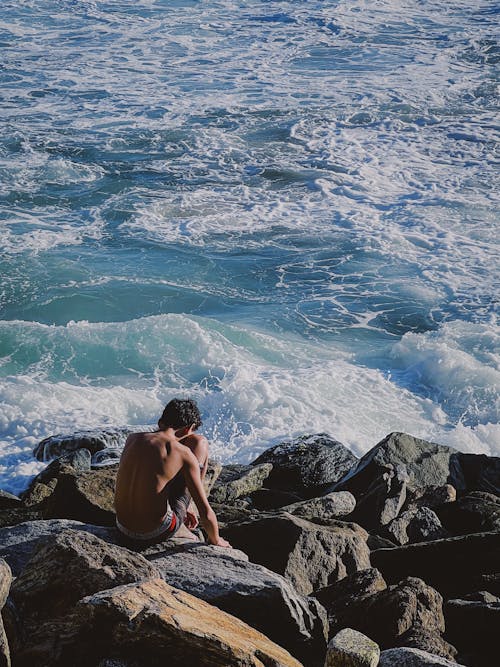 A Shirtless Man Sitting on a Rocky Shore