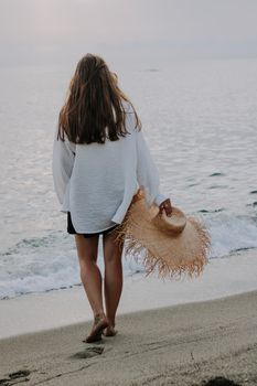 A woman walks barefoot on a sandy beach, holding a straw hat, during a peaceful sunset by the ocean.