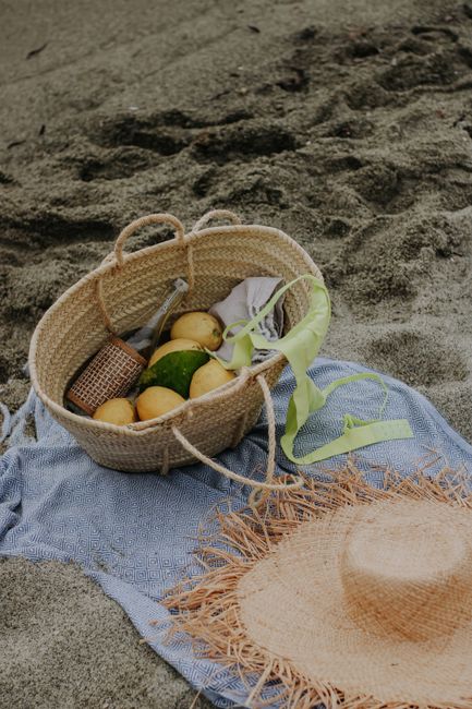 Woven beach basket with fruits and straw hat on a sandy blanket, perfect beach day setup.