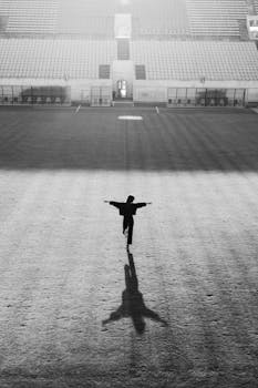 Black and white silhouette of a person standing with arms outstretched in a stadium.