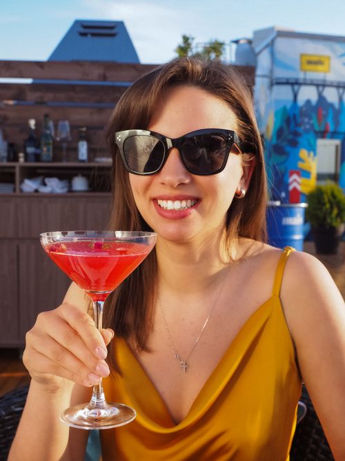 Free Smiling woman holding a cocktail outside in sunny weather, wearing sunglasses and a yellow dress. Stock Photo