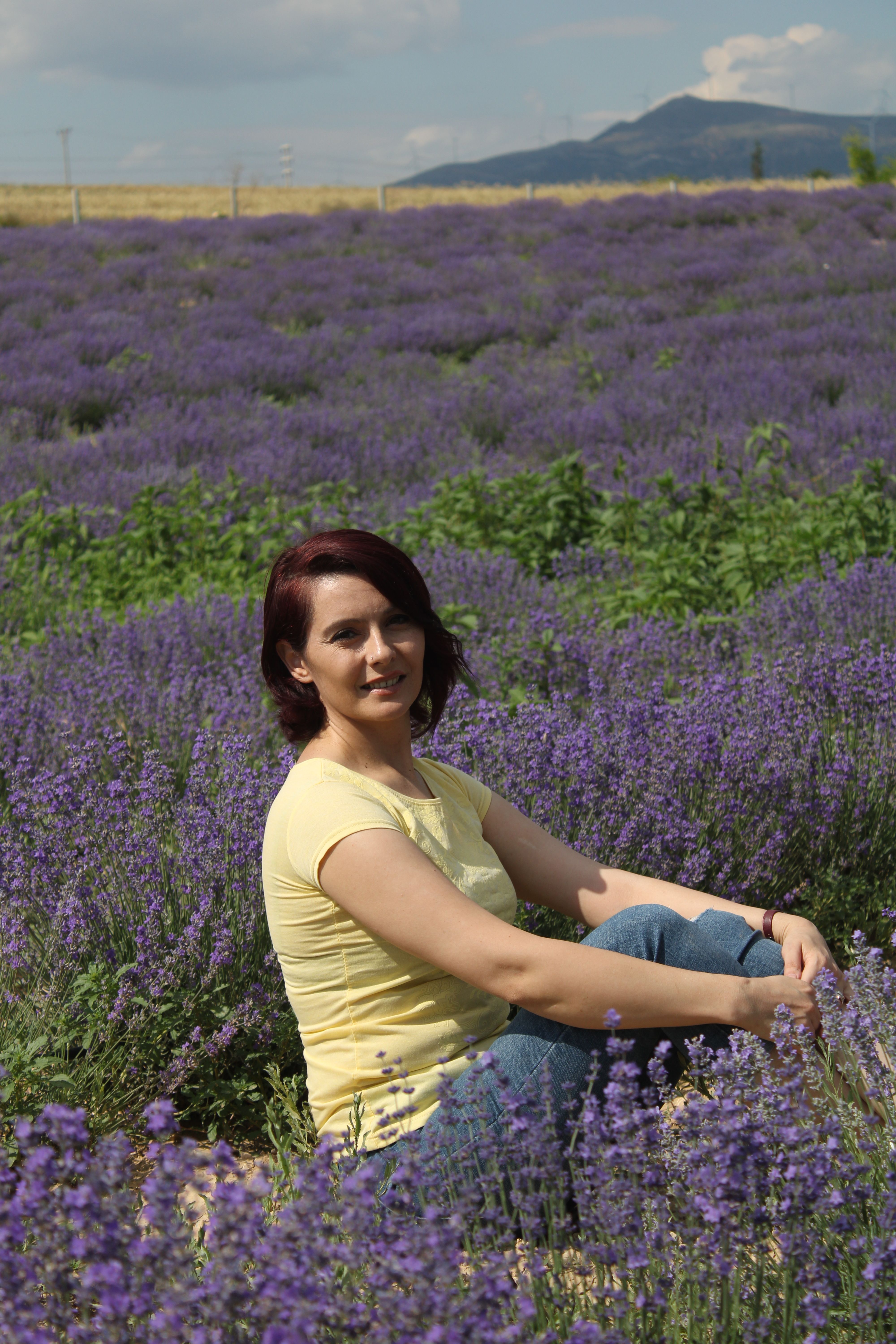 A woman sits smiling in a vibrant lavender field under a sunny sky.