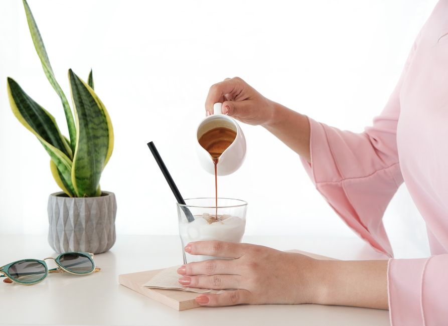 A woman pours coffee into a milk-filled glass cup with a straw, next to a plant and sunglasses indoors.