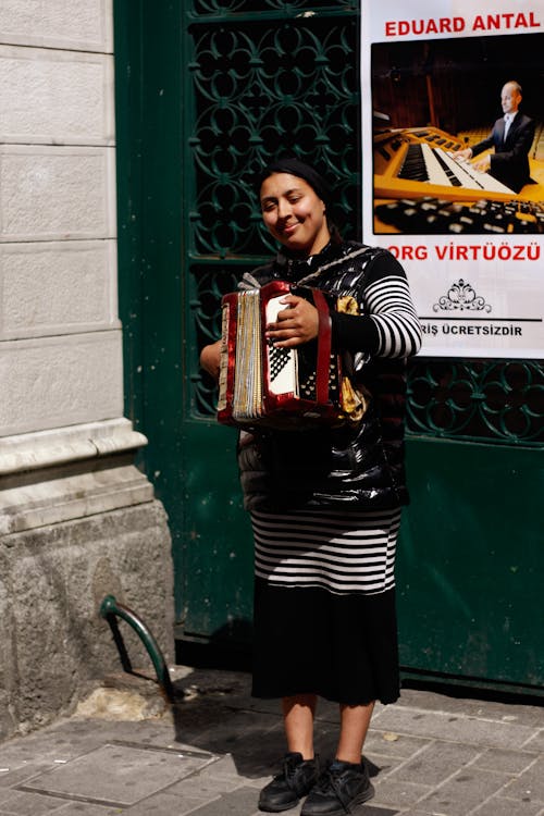 A Woman in Striped Long Sleeve Dress Playing Accordion while Standing on the Street