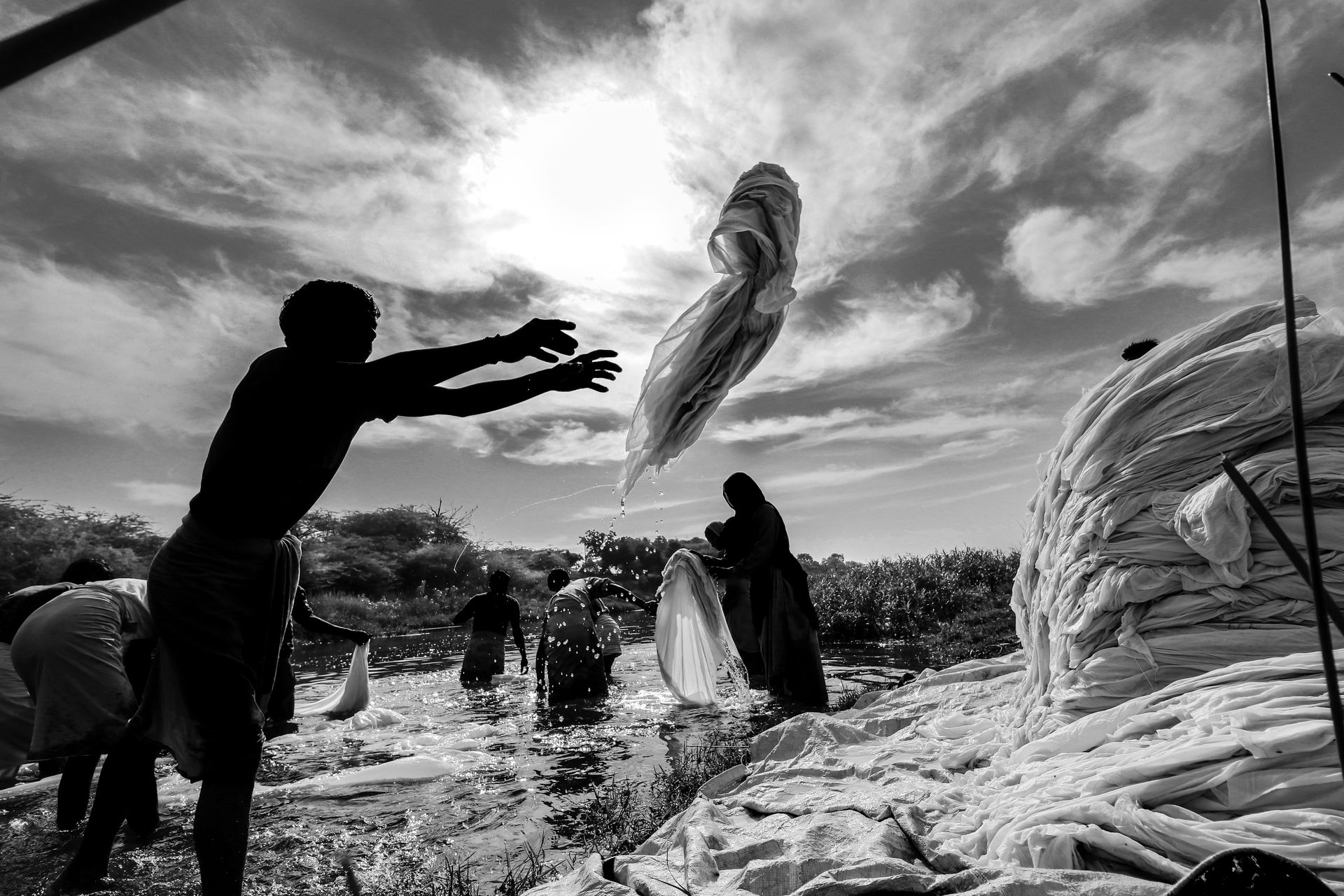 Silhouetted people washing textiles outdoors in Bagh, India, under a bright sky.
