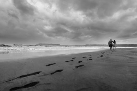 Monochrome image of a couple walking along Vagator Beach, Goa, with dramatic clouds and ocean waves.